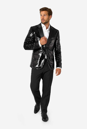 Man in a black sequin blazer with satin lapels and black dress pants, walking forward with one hand on his chest while looking to the side.