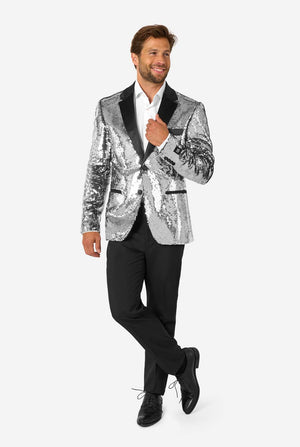 Full-body view of a man in a silver sequin blazer with black satin lapel, smiling with one hand near the jacket button.