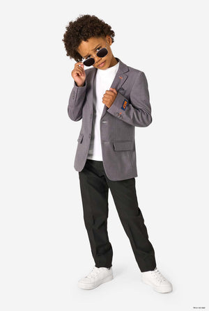 A young boy wearing a grey blazer, black trousers, and sunglasses, posing confidently while looking over the frames.