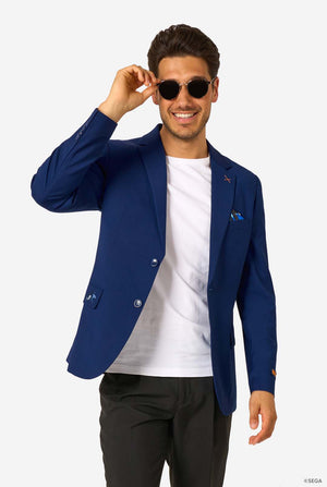 A man wearing a blue blazer, white T-shirt, black trousers, and sunglasses, smiling while lifting his shades playfully.