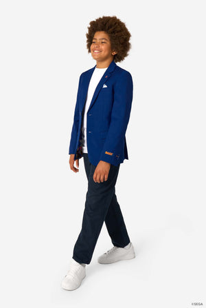 A child walking confidently in a blue blazer, white T-shirt, dark trousers, and white sneakers, smiling.