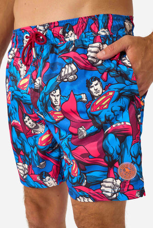 Man wearing a Men's Superman-themed swim shorts featuring a bold comic book design with multiple action poses of the Man of Steel. Perfect for superhero fans looking to make a splash. Close up front.