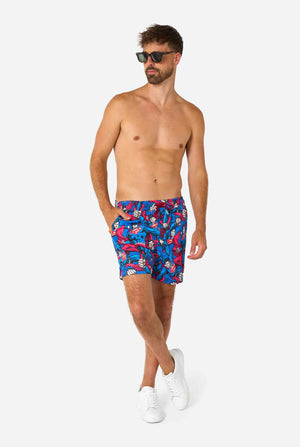 Man wearing a Men's Superman-themed swim shorts featuring a bold comic book design with multiple action poses of the Man of Steel. Perfect for superhero fans looking to make a splash. Full body image with man wearing sunglasses.