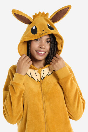 A woman is wearing an adorable Eevee onesie, modeled after the beloved Pokémon character. This adult unisex onesie is designed in a warm brown color, resembling Eevee’s fur, and features a fluffy beige collar mimicking Eevee’s mane. The hood includes Eevee's signature large brown ears, big black eyes. The onesie has a front zipper closure and convenient pockets at the front, offering both comfort and ease of wear. View of the upper part and holding the hood with two hands.
