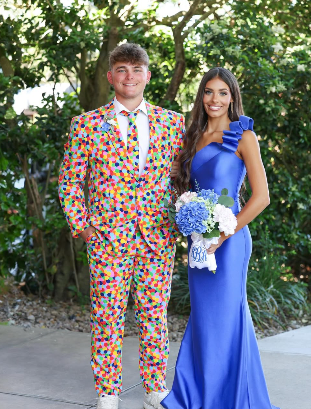 Image of a man and a woman. The woman is wearing a purple dress and the man is wearing a confetteroni suit.