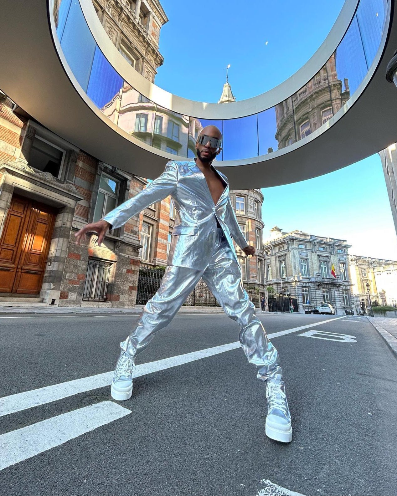 Person in a silver reflective suit posing on a city street with a modern building in the background.