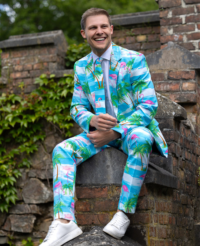 Man sitting outside on castle wall, wearing light blue suit with palm tree and flamingo print. Also wearing white shirts and white sneakers.