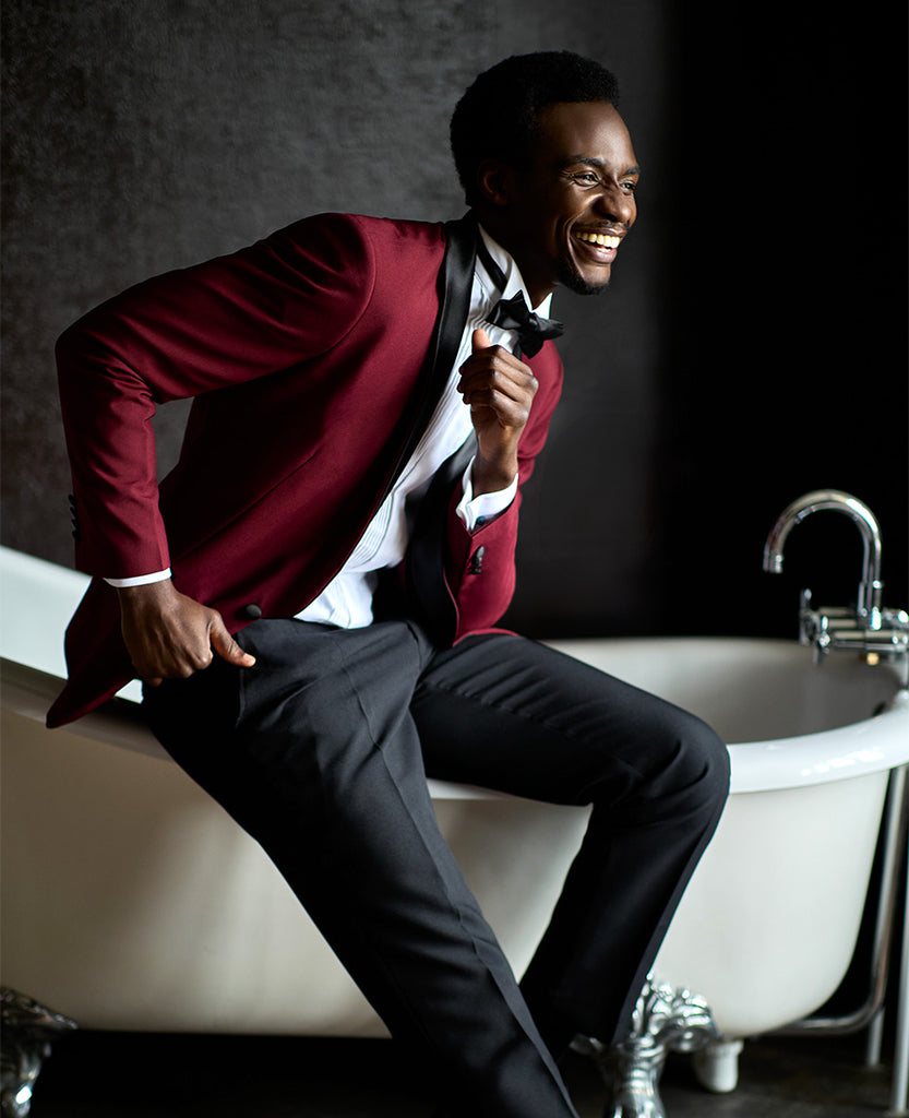 Man sitting on bathtub wearing tuxedo with burgundy red jacket, black pants an black bow tie. Also wearing white shirt.