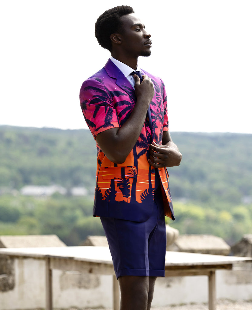 Man standing outdoors wearing summer suit with palm trees on the jacket. Pants is dark Blue and jacket fades from purple at the top to orange at the bottom with a dark blue border.