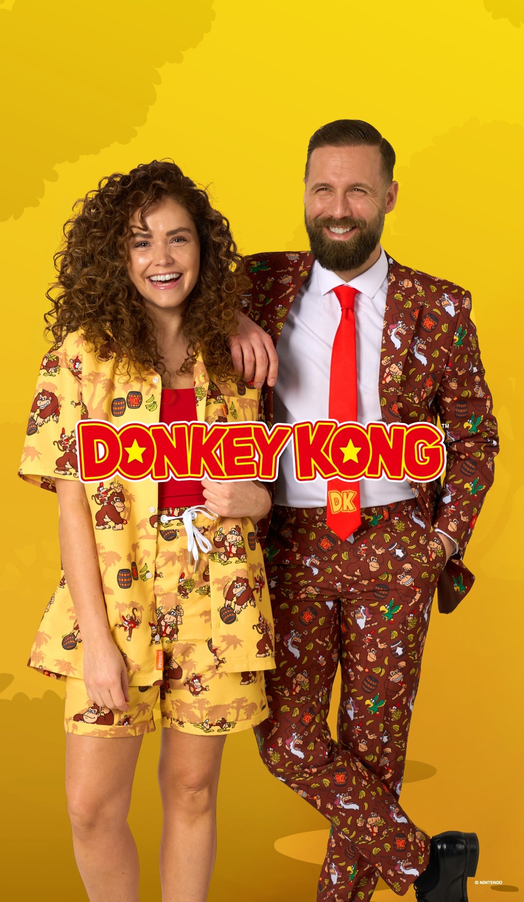 A man and a woman posing together in matching Donkey Kong themed outfits against a bright yellow background. The woman wears a yellow patterned pajama set, and the man wears a brown patterned suit with a red tie.