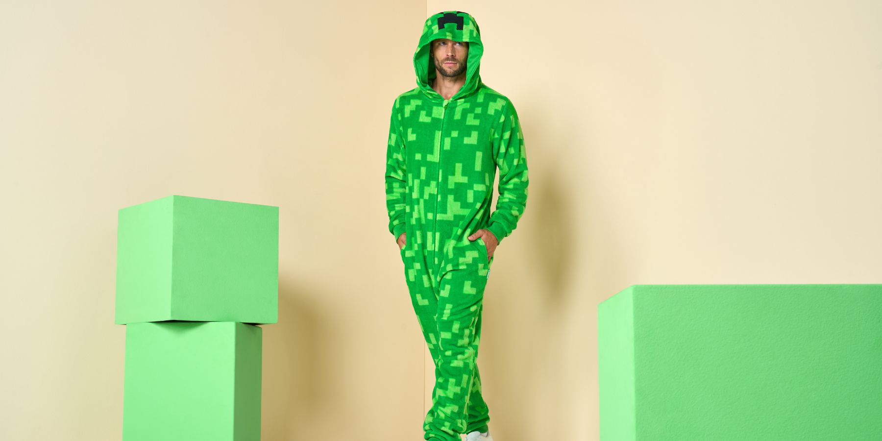 A man wearing a bright green pixelated Minecraft-style hooded onesie stands between two light green block props against a beige studio background.
