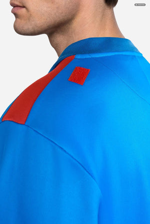 Close-up side view of a person wearing a bright blue knit top with a red shoulder panel and a small red square patch near the collar, against a light gray background.