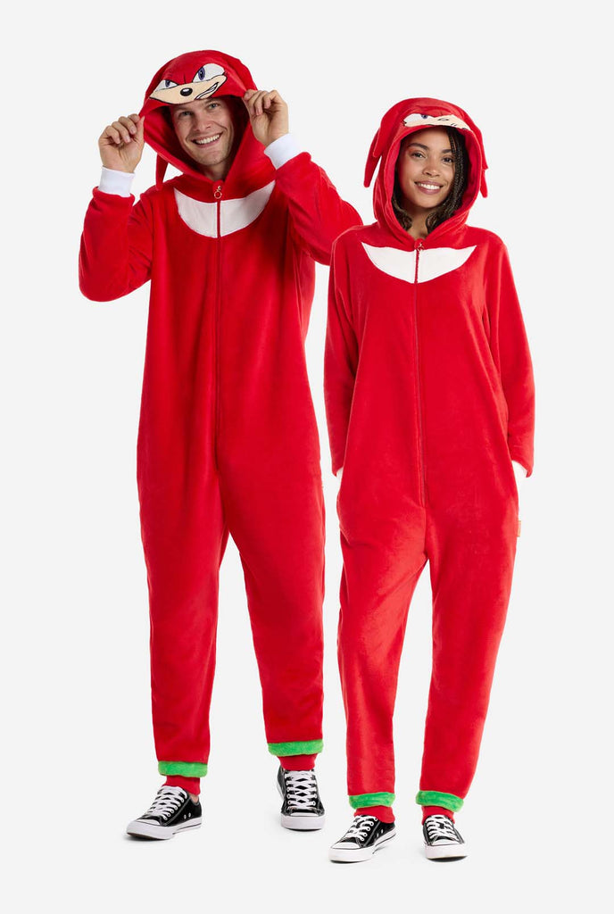 A man and woman are wearing a bright red Knuckles the Echidna-themed adult onesie from OppoSuits. The fun and cozy onesie features a zip-up front and an attached hood designed to resemble Knuckles' face, complete with expressive eyes and floppy dreadlocks. The suit is primarily red, with a white crescent pattern on the chest, reflecting Knuckles' iconic look from the Sonic the Hedgehog video game series. Full body image.