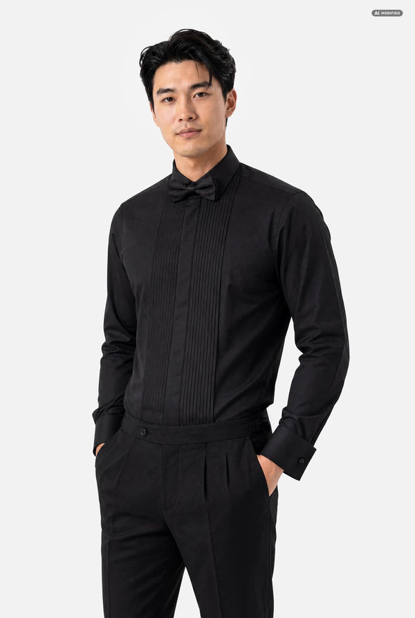 A man wearing a black long-sleeve pleated tuxedo shirt with a matching black bow tie and black trousers, standing against a light gray studio background.