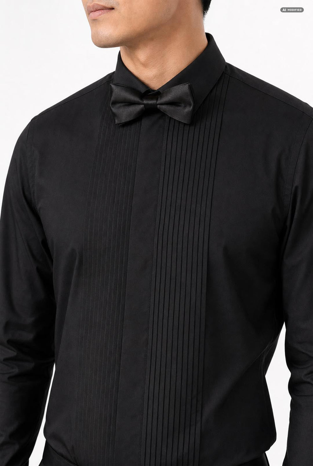 A man wearing a black long-sleeve pleated tuxedo shirt with a matching black bow tie and black trousers, standing against a light gray studio background.