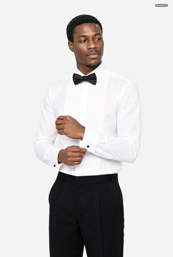 A man wearing a white long-sleeve tuxedo shirt with black bow tie and black trousers, adjusting his cuffs against a light gray studio background.