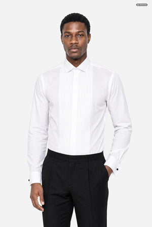 A man wearing a white long-sleeve tuxedo shirt with vertical pleats on the front and black trousers, standing against a light gray studio background.
