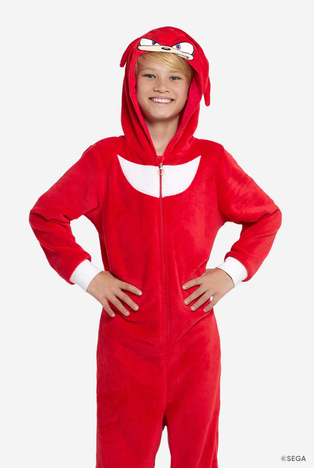 Smiling child standing front-facing, wearing a red Knuckles onesie with hood up and hands on hips. The onesie features a white chest patch and animated face on the hood.