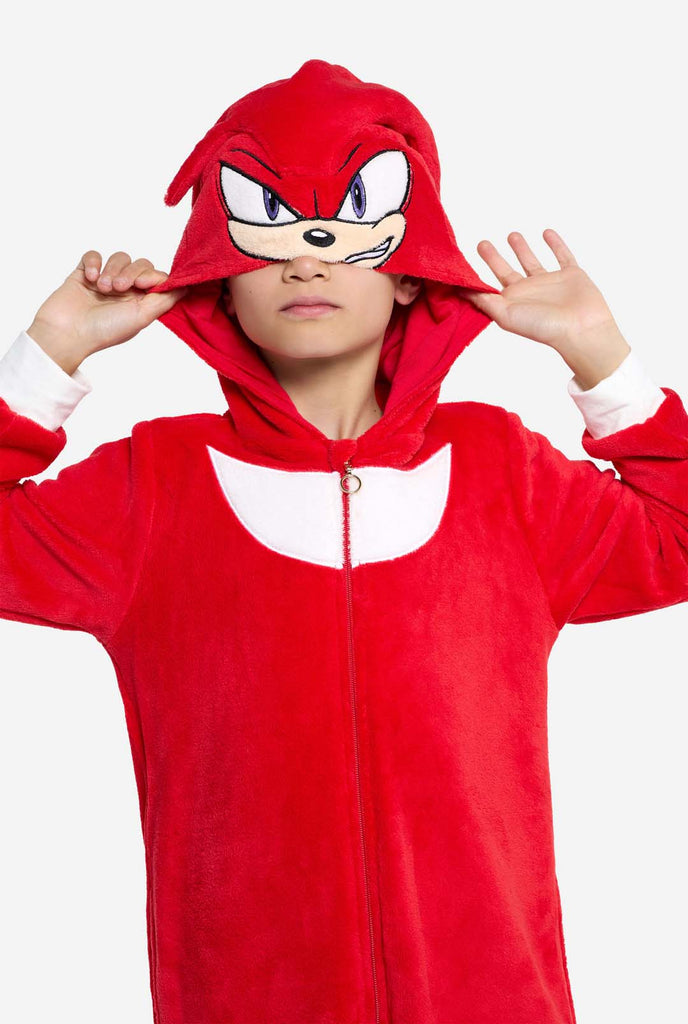 A kid is wearing a bright red Knuckles the Echidna-themed kids onesie from OppoSuits. The fun and cozy onesie features a zip-up front and an attached hood designed to resemble Knuckles' face, complete with expressive eyes and floppy dreadlocks. The suit is primarily red, with a white crescent pattern on the chest, reflecting Knuckles' iconic look from the Sonic the Hedgehog video game series. Holding hood over his eyes.