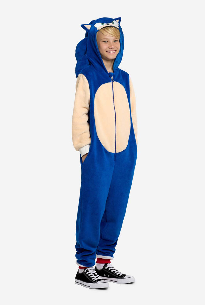 A kid is wearing a playful blue and beige Sonic the Hedgehog-themed kid onesie from OppoSuits. The cozy onesie features a zip-up front and an attached hood designed to resemble Sonic’s iconic face, complete with ears and an expressive gaze. The blue body of the suit contrasts with a beige belly, mimicking Sonic's classic look. This fun, comfortable, and stylish onesie is perfect for lounging, themed parties, or cosplay events, bringing nostalgic video game vibes to any occasion. Full body image.