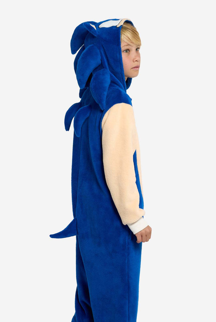 A kid is wearing a playful blue and beige Sonic the Hedgehog-themed kid onesie from OppoSuits. The cozy onesie features a zip-up front and an attached hood designed to resemble Sonic’s iconic face, complete with ears and an expressive gaze. The blue body of the suit contrasts with a beige belly, mimicking Sonic's classic look. This fun, comfortable, and stylish onesie is perfect for lounging, themed parties, or cosplay events, bringing nostalgic video game vibes to any occasion. Side view.