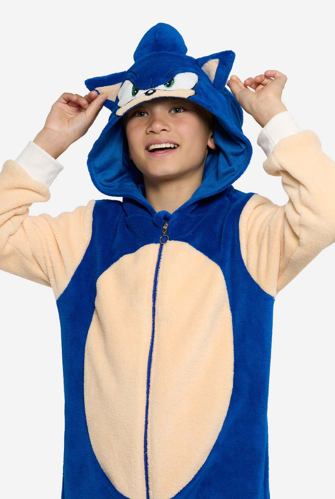 A kid is wearing a playful blue and beige Sonic the Hedgehog-themed kid onesie from OppoSuits. The cozy onesie features a zip-up front and an attached hood designed to resemble Sonic’s iconic face, complete with ears and an expressive gaze. The blue body of the suit contrasts with a beige belly, mimicking Sonic's classic look. This fun, comfortable, and stylish onesie is perfect for lounging, themed parties, or cosplay events, bringing nostalgic video game vibes to any occasion. Kid holding the hood