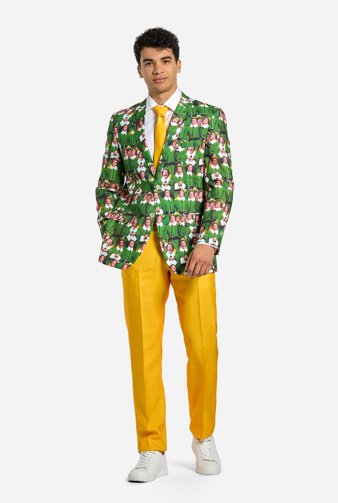 Man wearing men's Christmas suit with the character ELF on the jacket and yellow pants. Suits consists of all over print jacket with ELF Christmas movie character, yellow pants and yellow tie. Man also wearing a white shirt and white sneakers.