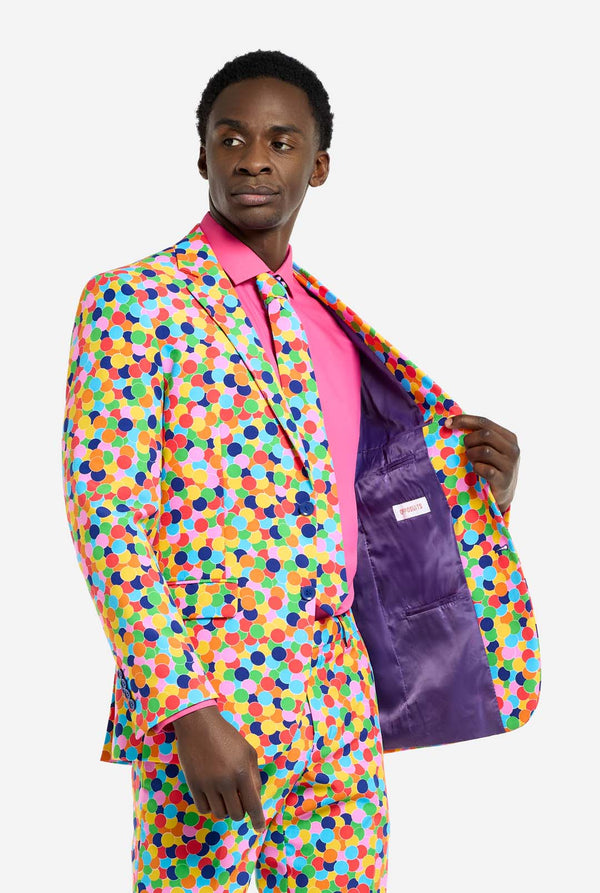 Man wearing men's suit with multicolor all-over confetti print and pink dress shirt. Suit consist of jacket, pants and tie. Holding one side of the jacket open.