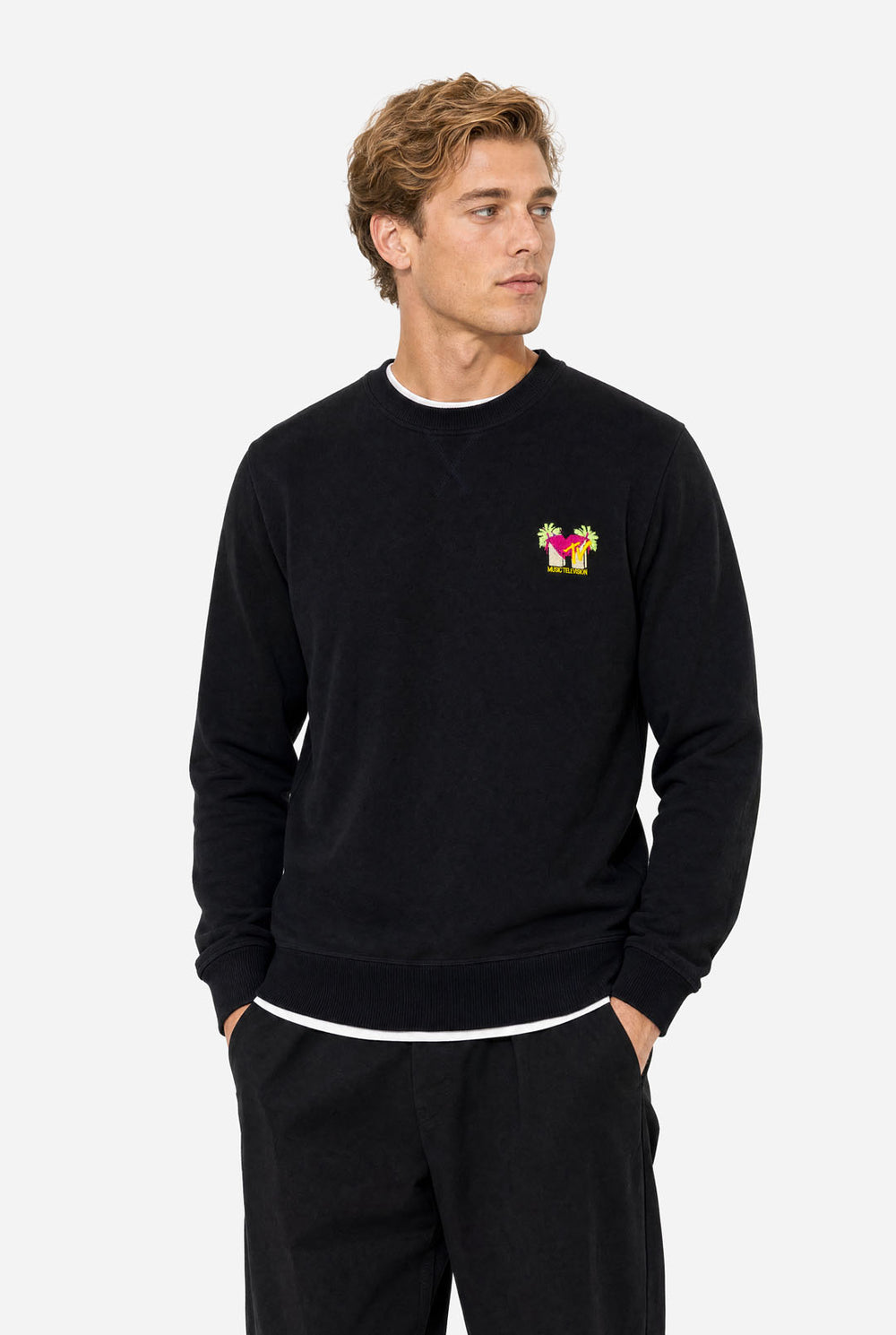 Man wearing a black crewneck sweatshirt against a light gray background, featuring a MTV logo embroidery in Pink and yellow on the left chest.