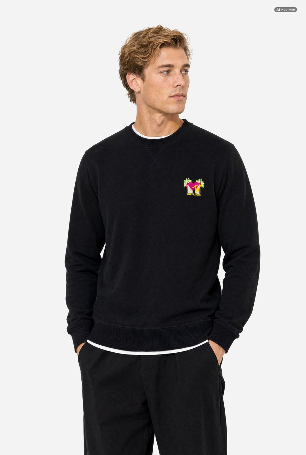 A man wearing a black crewneck sweatshirt with a small colorful embroidered MTV graphic on the left chest, paired with black sweatpants, against a light gray background.