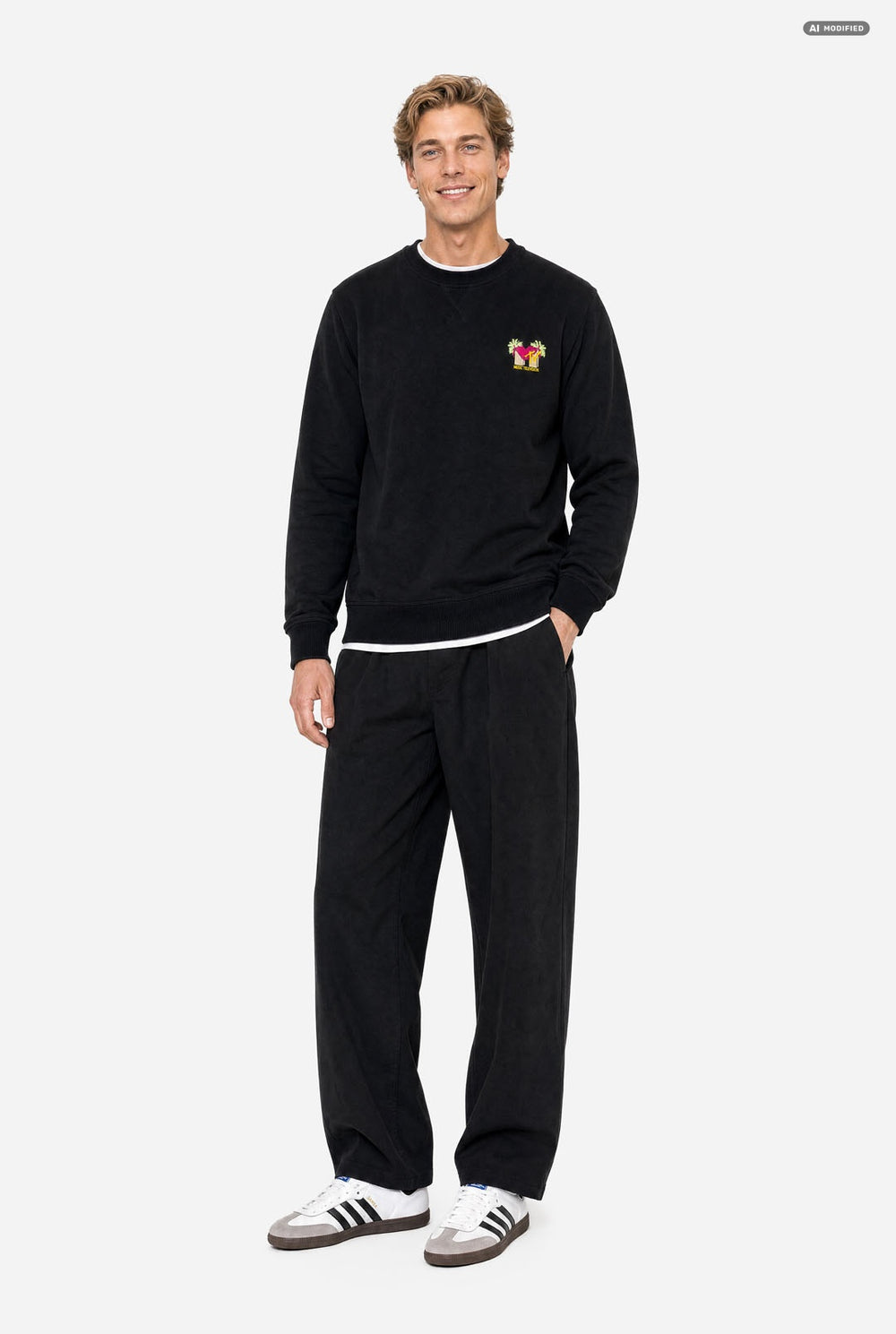 A man wearing a black crewneck sweatshirt with a small colorful embroidered MTV graphic on the left chest, paired with black sweatpants, against a light gray background.