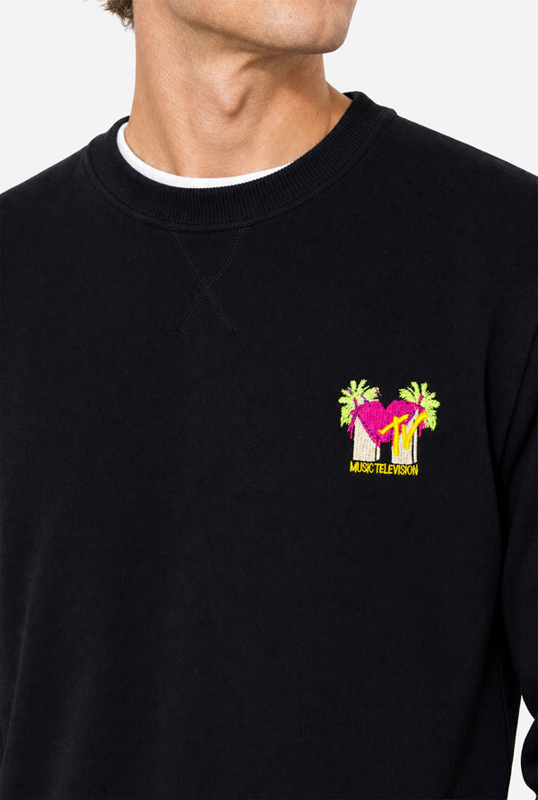 Man wearing a black crewneck sweatshirt against a light gray background, featuring a MTV logo embroidery in Pink and yellow on the left chest. Close up.