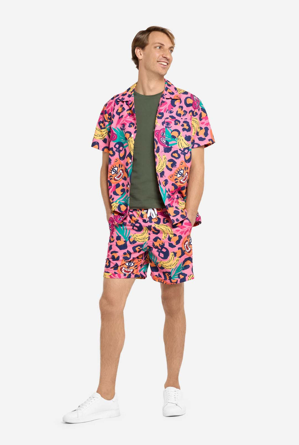 A man wearing a vibrant two-piece summer outfit featuring a bold tropical print. The short-sleeve button-up shirt and matching shorts showcase a lively design with neon pink, orange, and yellow leopard prints, bananas, green palm leaves, and quirky cartoon faces. The outfit is styled casually with a green T-shirt underneath and is perfect for summer parties, vacations, or poolside fun. 