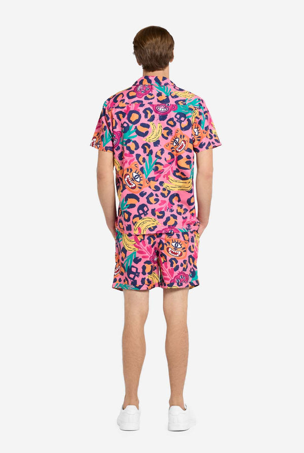 A man wearing a vibrant two-piece summer outfit featuring a bold tropical print. The short-sleeve button-up shirt and matching shorts showcase a lively design with neon pink, orange, and yellow leopard prints, bananas, green palm leaves, and quirky cartoon faces. The outfit is styled casually with a green T-shirt underneath and is perfect for summer parties, vacations, or poolside fun. View from the back.