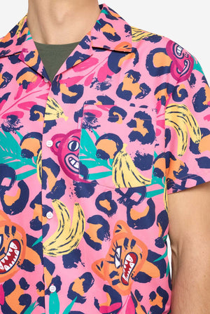 A man wearing a vibrant two-piece summer outfit featuring a bold tropical print. The short-sleeve button-up shirt and matching shorts showcase a lively design with neon pink, orange, and yellow leopard prints, bananas, green palm leaves, and quirky cartoon faces. The outfit is styled casually with a green T-shirt underneath and is perfect for summer parties, vacations, or poolside fun. Chest close up.