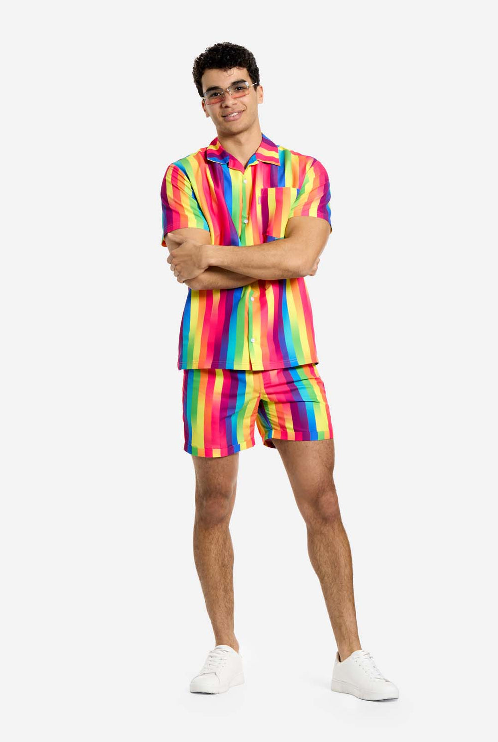 A man is wearing a vertically striped rainbow-colored summer set, consisting of shirt and shorts. He is smiling in the camera and holding one hand in his pocket.