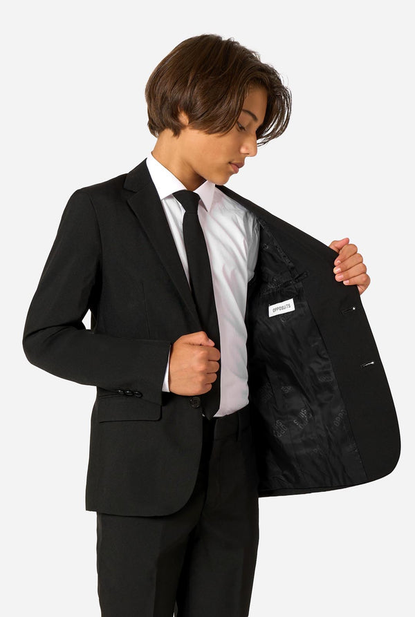 Teenager wearing a black suit and a white shirt on a light background. Holding one side of the jacket open.