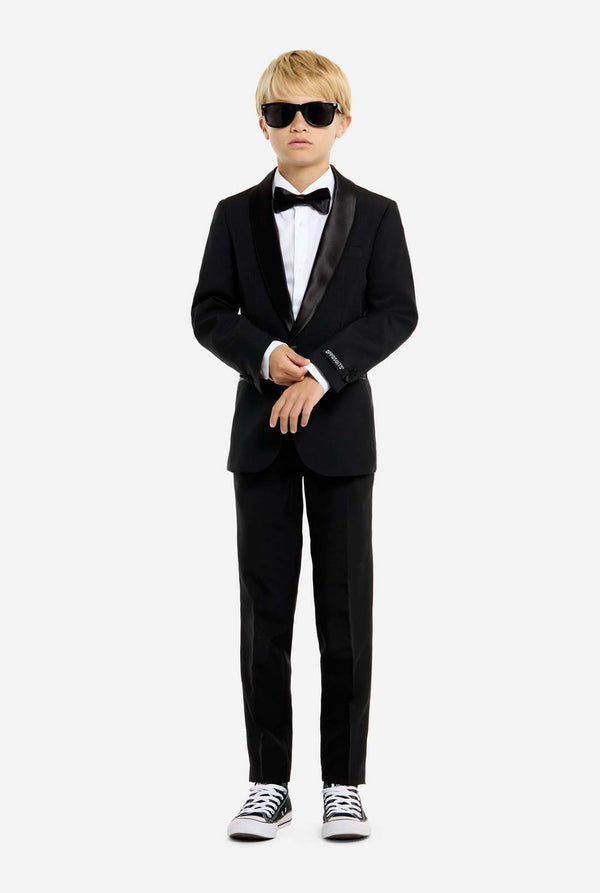 Person wearing a black tuxedo with sunglasses and white sneakers on a white background