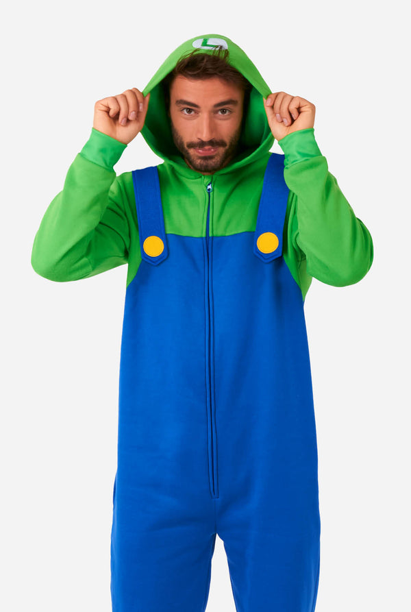A man is dressed in a Luigi-themed onesie. The outfit look as a bright green hoodie with an "L" symbol on the hood and blue overalls with large yellow buttons on the straps. He is holding both sides of the hood, as if pulling it over his head, and looking directly at the camera with a neutral expression. The overall design mimics the look of the popular video game character Luigi, with contrasting green and blue colors. The background is plain, putting focus on the costume and the man's playful pose.