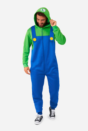 A man is dressed in a Luigi-themed onesie. The outfit look as a bright green hoodie with an "L" symbol on the hood and blue overalls with large yellow buttons on the straps. He is holding one side of the hood, as if pulling it over his head, and looking at the floor. The overall design mimics the look of the popular video game character Luigi, with contrasting green and blue colors. The background is plain, putting focus on the costume and the man's playful pose.