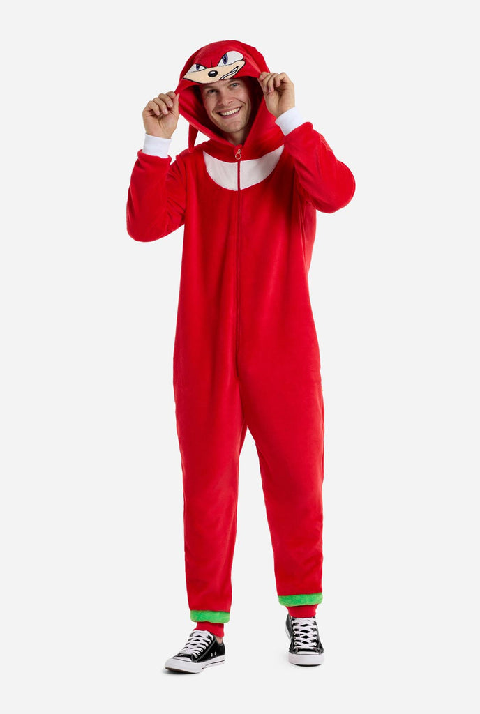 A man is wearing a bright red Knuckles the Echidna-themed adult onesie from OppoSuits. The fun and cozy onesie features a zip-up front and an attached hood designed to resemble Knuckles' face, complete with expressive eyes and floppy dreadlocks. The suit is primarily red, with a white crescent pattern on the chest, reflecting Knuckles' iconic look from the Sonic the Hedgehog video game series. Full body image.