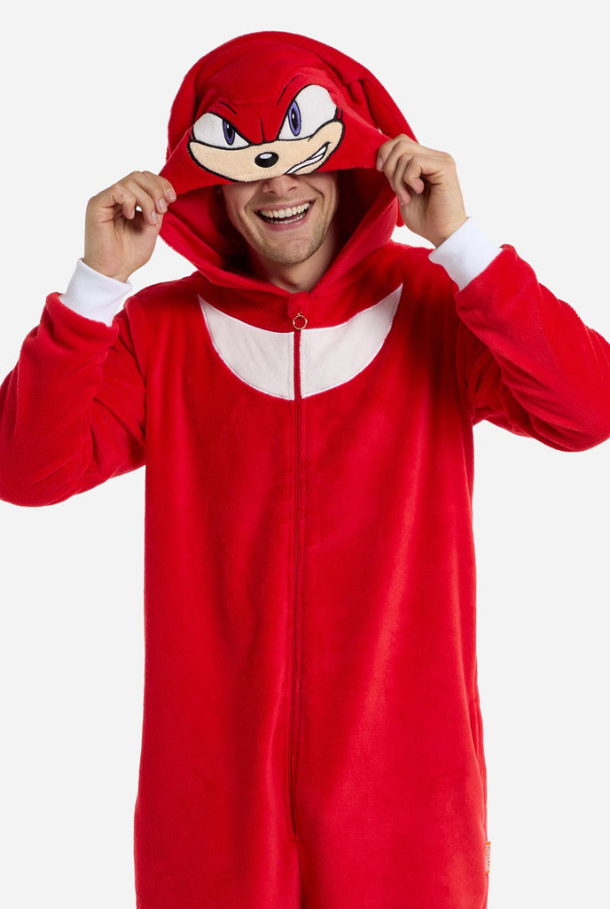 A man is wearing a bright red Knuckles the Echidna-themed adult onesie from OppoSuits. The fun and cozy onesie features a zip-up front and an attached hood designed to resemble Knuckles' face, complete with expressive eyes and floppy dreadlocks. The suit is primarily red, with a white crescent pattern on the chest, reflecting Knuckles' iconic look from the Sonic the Hedgehog video game series. The man is holding the hood over his eyes.