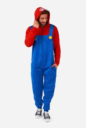 A man is dressed in a Mario-themed onesie. The outfit look as a bright red hoodie with an "M" symbol on the hood and blue overalls with large yellow buttons on the straps. He is holding one side of the hood, as if pulling it over his head, and looking sideways at the floor. The overall design mimics the look of the popular video game character Mario, with contrasting red and blue colors. The background is plain, putting focus on the costume and the man's playful pose.