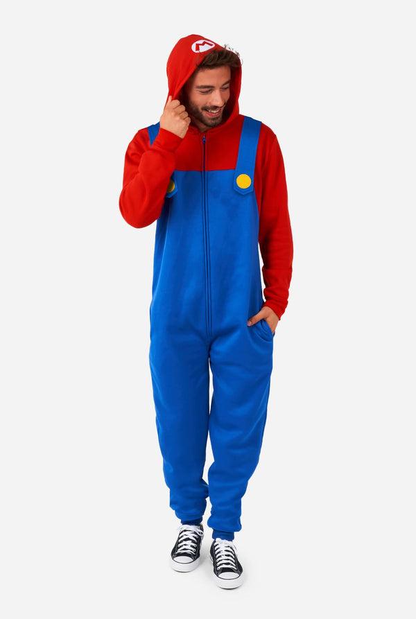 A man is dressed in a Mario-themed onesie. The outfit look as a bright red hoodie with an "M" symbol on the hood and blue overalls with large yellow buttons on the straps. He is holding one side of the hood, as if pulling it over his head, and looking sideways at the floor. The overall design mimics the look of the popular video game character Mario, with contrasting red and blue colors. The background is plain, putting focus on the costume and the man's playful pose.