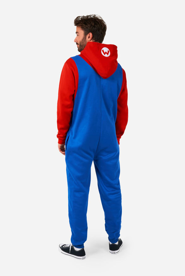 A man is dressed in a Mario-themed onesie. The outfit looks as a bright red hoodie with an "M" symbol on the hood and blue overalls with large yellow buttons on the straps. View from the back and the hood flat on the back. The overall design mimics the look of the popular video game character Mario, with contrasting red and blue colors. The background is plain, putting focus on the costume.