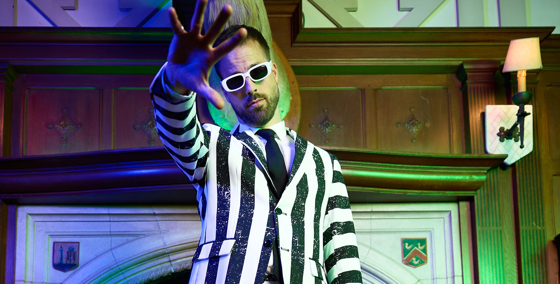 A man wearing a black and white vertically striped suit from the movie Beetlejuice, a dark tie, and white sunglasses poses with his hand raised in front of a decorative wooden interior wall.