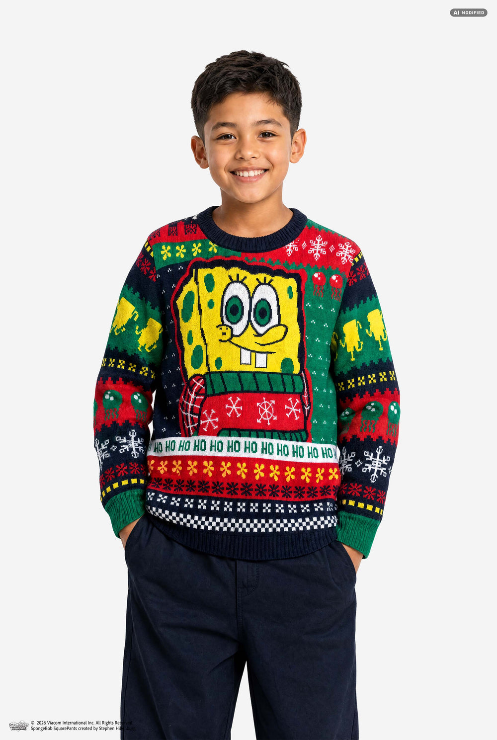 A smiling young boy wearing a festive SpongeBob-themed Christmas sweater and dark navy pants, standing against a plain light gray studio background.