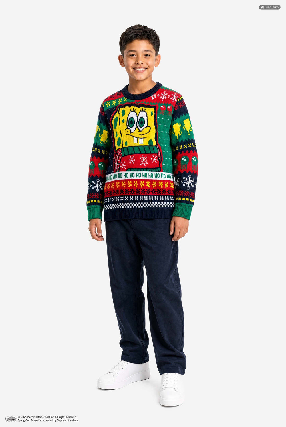 A smiling young boy wearing a festive SpongeBob-themed Christmas sweater and dark navy pants, standing against a plain light gray studio background.