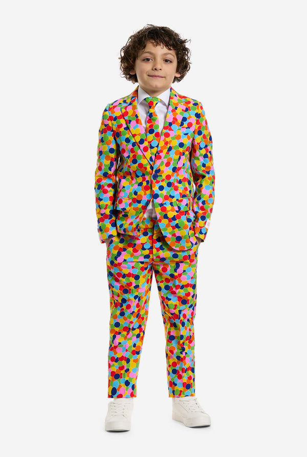 In this image, a kid is wearing a boys’ suit with all-over multicolor confetti print and white shirt. The suit has a bright pattern with circles in various colors including orange, yellow, pink, green, blue, and red. Kid has hands in his pockets.