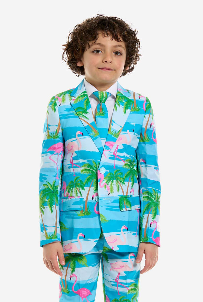 In this image, a kid is wearing a light blue boys’ suit with a tropical print and a white shirt. The suit has a blue print resembling water and pink flamingos and green palm trees.